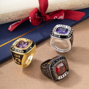 May include: Three class rings in gold, silver and black with different coloured gemstones. The rings feature the text "Class of" and the year "2023".