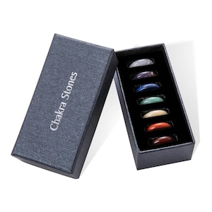 Może przedstawiać: Zestaw siedmiu kamieni czakr w niebieskim pudełku. Kamienie są ułożone w rzędzie i oznaczone odpowiadającymi im kolorami czakr. Pudełko jest oznaczone napisem "Chakra Stones".