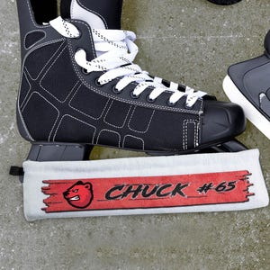 Puede incluir: Patín de hockey blanco y negro con una cubierta de cuchilla de patín de hockey blanca y roja. La cubierta tiene un logotipo de oso rojo y el texto "CHUCK #65".