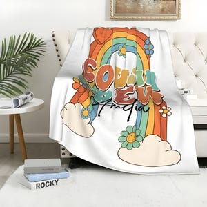 Op de afbeelding: Een wit fleece deken met een regenboog, wolken en de tekst "COULDN'T BEW" en "MEDIA" in een retro lettertype. De regenboog is gemaakt van rode, oranje, gele, groene, blauwe, indigo en violette strepen.