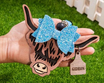 May include: A wooden Highland cow-shaped keychain with a light blue glitter bow. The cow has a brown body, black horns, and a tag that reads "GIDEON". The keychain is held against a green background.