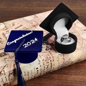 Może przedstawiać: Niebieski aksamitny czepek absolwenta z białym frędzlem i napisem "Congratulations! 2024" na górze. Czepek leży na książce, obok znajduje się pudełko na pierścionek. Pudełko na pierścionek jest z czarnego aksamitu i zawiera srebrny pierścionek.