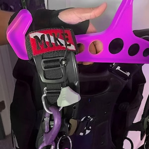 Puede incluir: Un chaleco de buceo negro con un colgador y accesorios morados. El chaleco tiene un parche que dice "MIKE" en letras blancas sobre un fondo rojo y blanco. El colgador y los accesorios son morados.