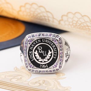 May include: A silver ring with a black enamel inlay featuring the words "Lock Haven University" and the initials "Bu" surrounded by a laurel wreath. The ring is set with purple gemstones and the year "1984" is inscribed below the wreath.