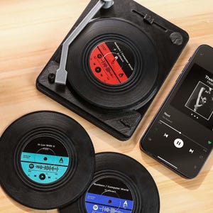 Puede incluir: Posavasos de vinilo negro con etiquetas coloridas y un tocadiscos en miniatura. Un smartphone muestra una interfaz de aplicación de música. Los posavasos presentan el texto "In Luv With U" y "Numbers / Computer World Kraftwerk".