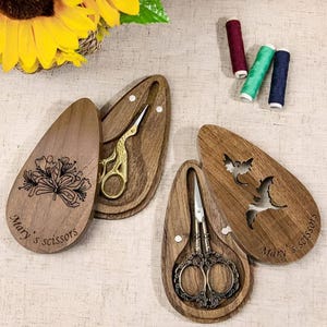 May include: Two wooden, teardrop-shaped cases with engraved floral and bird designs. Each case holds a pair of decorative scissors. The cases are labeled "Mary's Scissors".
