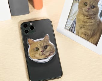 May include: A black smartphone with a clear cat-face phone grip. The grip features a close-up of a tan cat with green eyes. A framed photo of a similar cat is also visible.