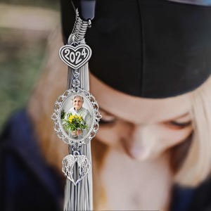 May include: A silver graduation tassel with a heart charm that says "2024" and a photo frame charm with a picture of a woman holding flowers. The tassel is hanging from a graduation cap.