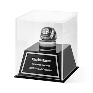 May include: A black and silver football championship ring displayed in a clear acrylic case. The ring sits atop a black base with a white framed plaque that reads "Chris Harm, Altaussee Cowboys, 2023 Football Champion."