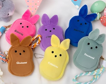 Op de afbeelding: Vijf pluche konijnenvormige muntbeursen in verschillende kleuren. De beursen zijn roze, paars, blauw, bruin en geel. Elke beurs heeft een andere naam geborduurd: Bonnie, Charlie, Bertram, Ferdinand en Colastio.