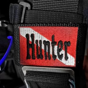 Op de afbeelding: Een zwarte duiktas met een rood-witte duikvlag patch met de tekst "Hunter" erop.