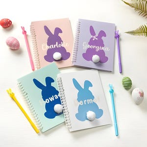 May include: Four spiral bound notebooks featuring bunny silhouettes and personalised names: Charlotte, Georgina, Owa, and Erm. Each notebook has a fluffy white pom-pom tail and comes with a colourful pen.