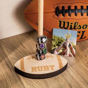 Puede incluir: Un soporte de anillos de madera con forma de balón de fútbol, con el nombre "RUBY" grabado. Muestra cinco anillos de campeonato con piedras preciosas de colores. Un balón de fútbol y una foto de un trofeo están en el fondo.