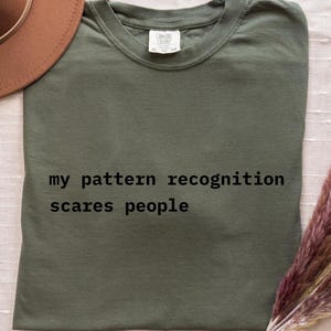 Op de afbeelding: Een olijfgroen t-shirt met de tekst "my pattern recognition scares people" in het zwart. Een bruine hoed en een bos gedroogd, roodbruin pampasgras staan ook op de afbeelding.
