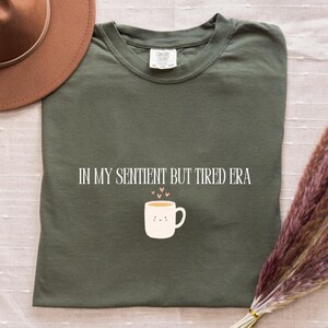 Op de afbeelding: Een olijfgroen t-shirt met de tekst "IN MY SENTIENT BUT TIRED ERA" en een afbeelding van een lachende koffiekop. Een bruine hoed en een bos gedroogd pampasgras staan ook op de afbeelding.