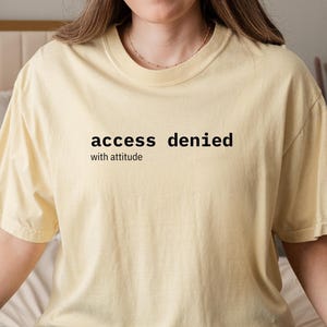 Op de afbeelding: Lichtgeel T-shirt met de tekst "access denied with attitude" in het zwart. Het shirt wordt gecombineerd met een blauwe spijkerbroek. De persoon zit op een bed met witte kussens en een beige dekbed.