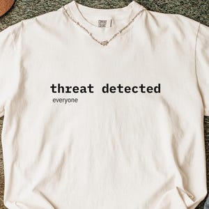 Op de afbeelding: Crèmekleurig T-shirt met de zwarte tekst "threat detected" en "everyone". De afbeelding toont witte sneakers, een bruine pet en haarclips. Een blauwe deken met witte harten is ook zichtbaar.