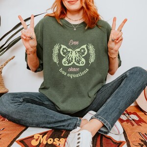 May include: A green t-shirt with a butterfly graphic and the text "Even chaos has equations". The model is wearing blue jeans and white sneakers.