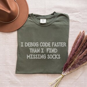 Op de afbeelding: Een olijfgroen t-shirt met de tekst "I debug code faster than I find missing socks" in het wit. Een bruine hoed en een bos gedroogd pampasgras staan ook op de foto.