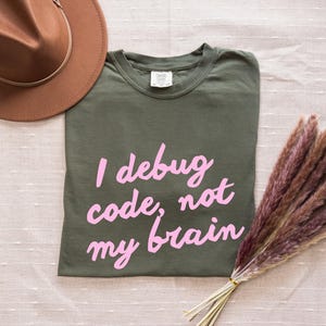 Op de afbeelding: Een olijfgroen t-shirt met de tekst "I debug code, not my brain" in roze letters. Een bruine hoed en een bos gedroogd, veervormig, bruin-paars pampasgras staan ook op de afbeelding.
