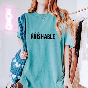 Op de afbeelding: Turquoisekleurig T-shirt met de zwarte tekst "you look PHISHABLE". Het shirt met korte mouwen is effen van kleur. De afbeelding toont ook een blauwe tas met witte hartvormen en een neonroze bord met de tekst "XOXO".