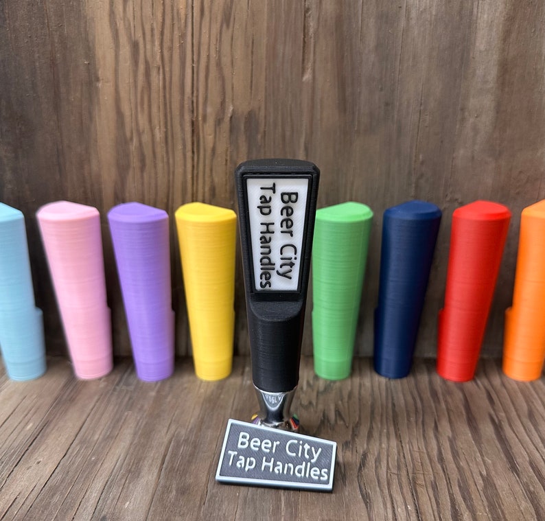 May include: Black tap handle with a white label that says "Beer City Tap Handles". The handle is surrounded by colorful tap handles in various colors, including blue, green, yellow, pink, and red.