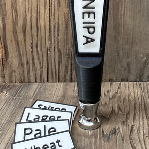 May include: A black beer tap handle with a white label that says "NEIPA". There are four white labels with black text on a wooden surface. The labels say "Saison", "Lager", "Pale", and "Wheat".