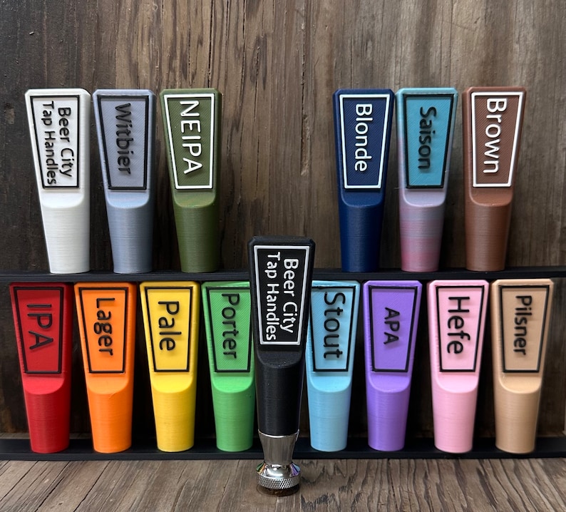 May include: A set of 12 colorful beer tap handles with different beer styles printed on them, including IPA, Pale Ale, Lager, Stout, and Pilsner. The handles are arranged in two rows on a black shelf.