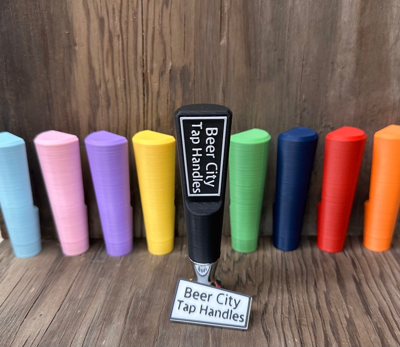 May include: A black tap handle with the text "Beer City Tap Handles" on a white label. The handle is surrounded by ten colorful tap handles in various colors, including blue, pink, purple, yellow, green, red, and orange.