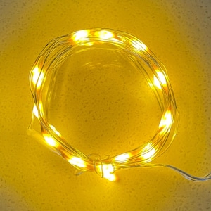 May include: A coiled string of warm white LED fairy lights. The lights are on a thin, flexible wire, creating a soft, ambient glow. The lights are arranged in a circular pattern, perfect for decorative purposes.