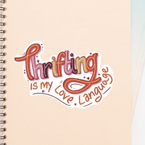 May include: A pink and beige notebook with a spiral binding. A sticker with the text "Thrifting is my love language" is on the front cover.
