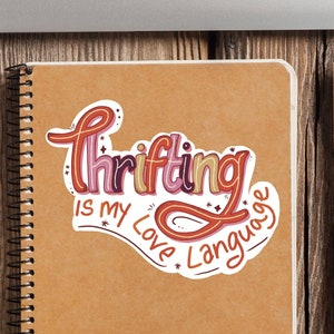 May include: A sticker with the text "Thrifting is my love language" in a colorful, handwritten font. The sticker is on a brown notebook with a spiral binding.