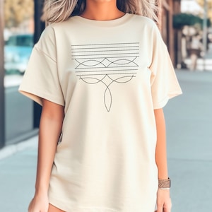 Puede incluir: Una camiseta beige de gran tama&ntilde;o con un dise&ntilde;o geom&eacute;trico negro en la parte delantera. El dise&ntilde;o presenta una serie de l&iacute;neas horizontales y formas curvas.