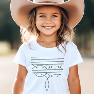 May include: A young girl wearing a white t-shirt with a black geometric design and a brown cowboy hat.