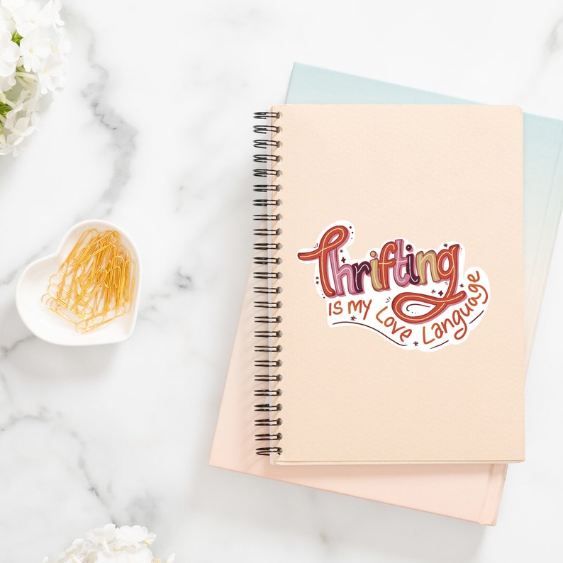 May include: A pink and beige notebook with a spiral binding. A sticker with the text "Thrifting is my love language" is on the front cover.