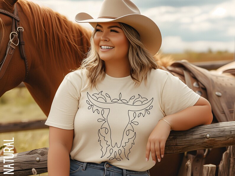 May include: A woman wearing a beige t-shirt with a black embroidered bull skull design. She is wearing a brown cowboy hat and blue jeans. She is standing next to a brown horse.