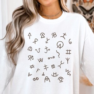 Op de afbeelding: Witte longsleeve sweatshirt met een bruine grafische afbeelding van verschillende symbolen en karakters die in een cirkel zijn gerangschikt.