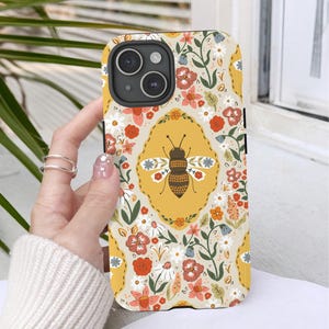 May include: A phone case with a floral and bee design. The case has a yellow background with a repeating pattern of flowers in red, orange, and white. A large bee is centered on the case, inside a yellow oval. The case is held by a hand.