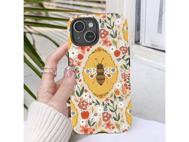 May include: A phone case with a floral and bee design. The case has a yellow background with a repeating pattern of flowers in red, orange, and white. A large bee is centered on the case, inside a yellow oval. The case is held by a hand.
