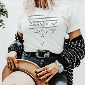 Puede incluir: Una camiseta blanca con un dise&ntilde;o geom&eacute;trico negro en la parte delantera. El dise&ntilde;o presenta una serie de l&iacute;neas horizontales y l&iacute;neas curvas que crean un patr&oacute;n sim&eacute;trico.