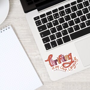 May include: A laptop computer with a sticker on the trackpad that says "Thrifting is my love language". The sticker is in a colorful, handwritten font.