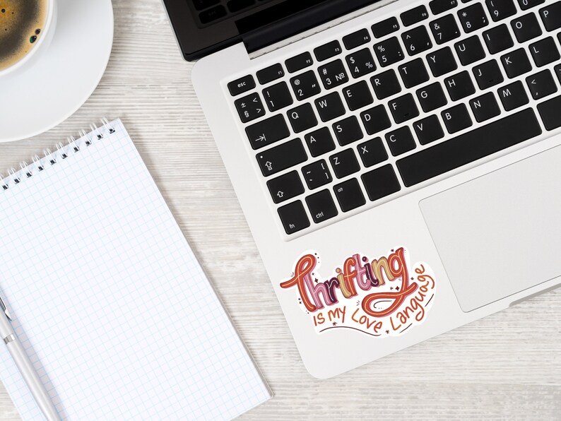 May include: A laptop computer with a sticker on the trackpad that says "Thrifting is my love language". The sticker is in a colorful, handwritten font.