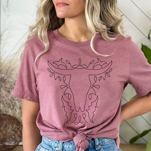 May include: A mauve t-shirt with a black embroidered design of a bull's head with floral accents.