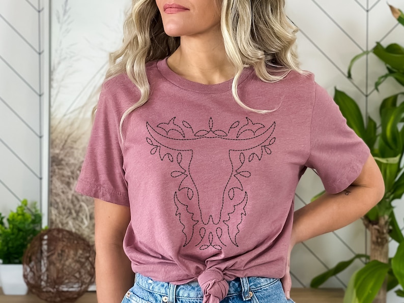 May include: A mauve t-shirt with a black embroidered design of a bull's head with floral accents.