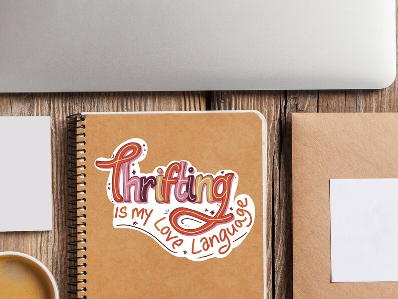 May include: A sticker with the text "Thrifting is my love language" in a colorful, handwritten font. The sticker is on a brown notebook with a spiral binding.