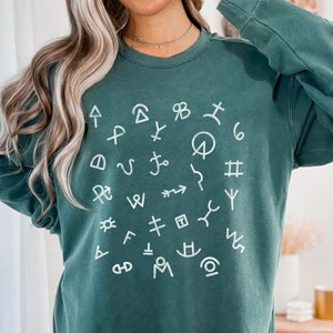 Op de afbeelding: Een tealgroen sweatshirt met een wit cirkelvormig ontwerp met verschillende symbolen en markeringen. Het sweatshirt heeft lange mouwen en een ronde hals.