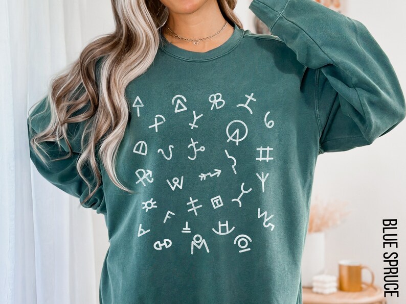 Op de afbeelding: Een tealgroen sweatshirt met een wit cirkelvormig ontwerp met verschillende symbolen en markeringen. Het sweatshirt heeft lange mouwen en een ronde hals.