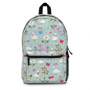 May include: A light blue backpack with a whimsical pattern of white daisies, strawberries, and geese wearing bows. The backpack has a black handle, a front pocket, and a side mesh pocket. The design includes pink and blue bows.