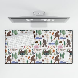 May include: A rectangular desk mat with a Bigfoot and nature-themed pattern. The design features Bigfoot figures, trees, mountains, cabins, and woodland creatures in a repeating pattern on a white background. A white keyboard and mouse are on the mat.