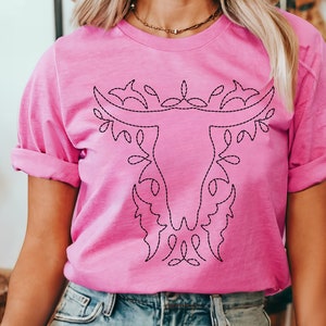 May include: A pink t-shirt with a black embroidered design of a stylized bull's head with floral accents. The text "HEATHER CHARITY PINK" is visible on the left side of the image.